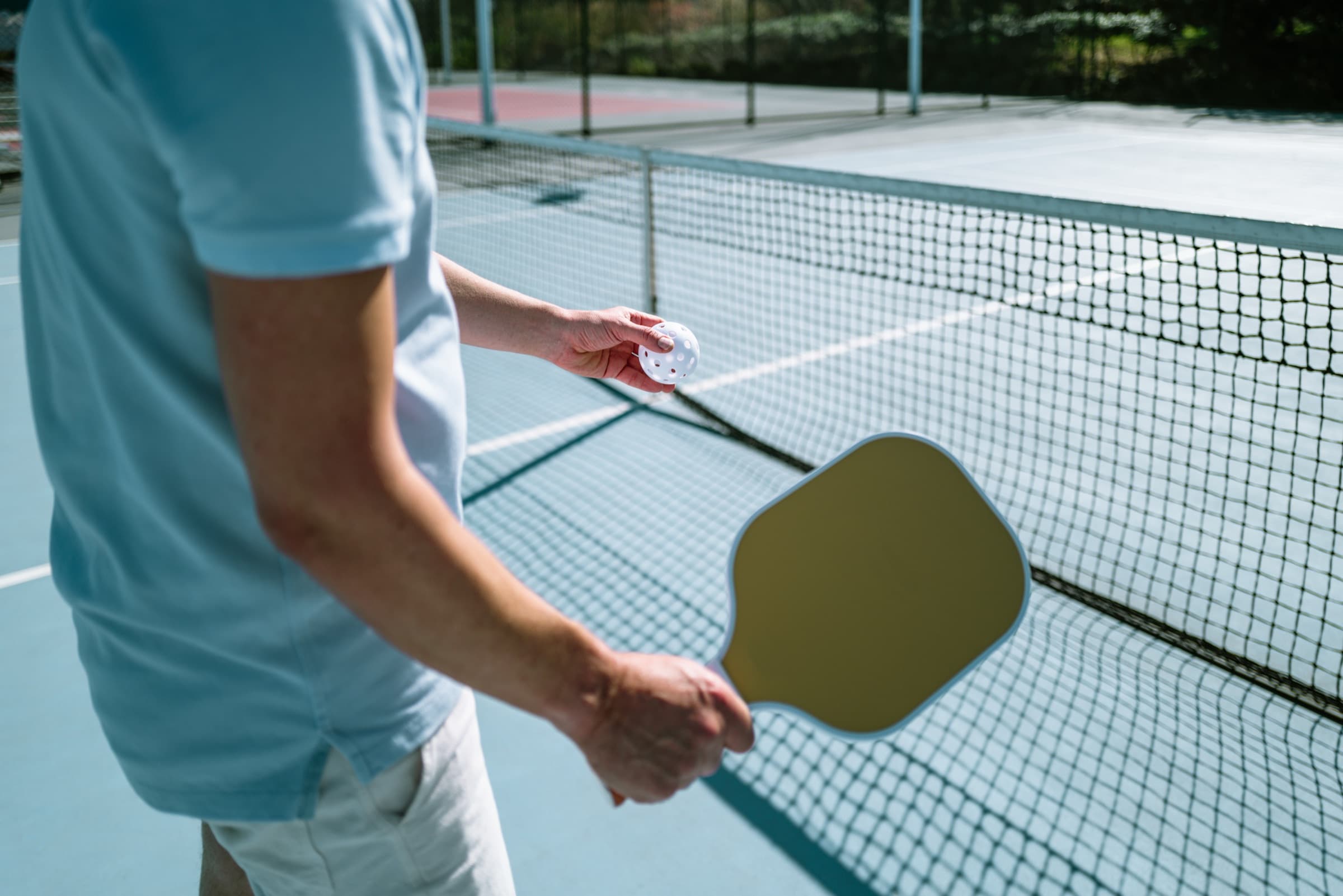 Player holding a pickleball paddle and ball at the net on an outdoor court